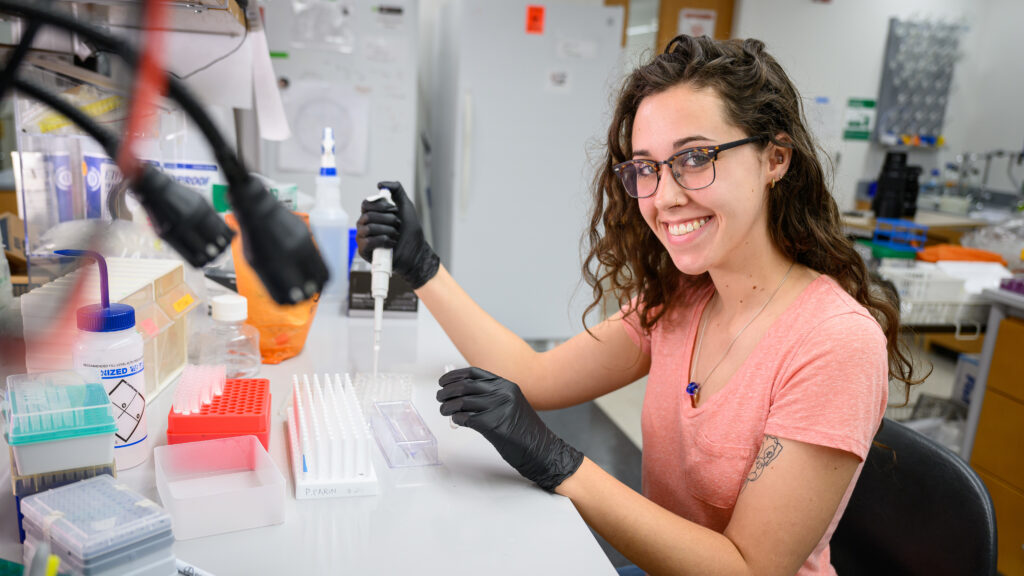 Meg Mulder, wearing a pink shirt and black gloves while sitting in a research lab, smiles at the camera while using a dropper to insert material into test tubes.
