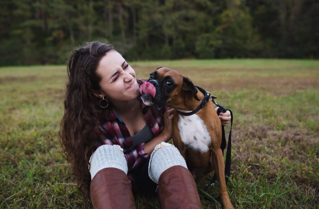 Meg Mulder sits on a grassy field in front of a line of trees with her dog, a brown boxer named Kona. She scrunches her face as Kona licks her chin and looks into the camera.