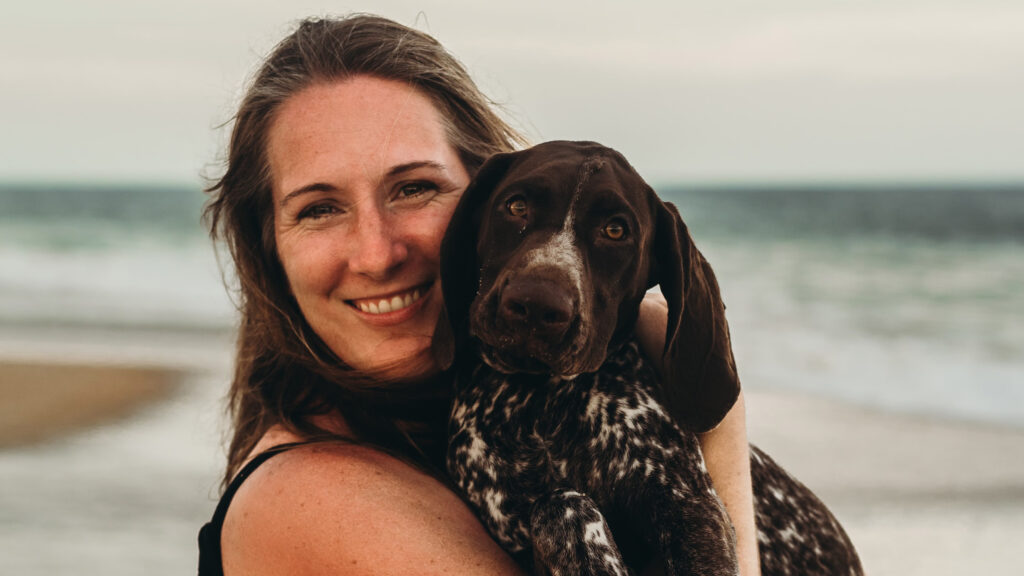 A brunette woman smiles at the camera while holding a brown-and-white puppy on a beach.