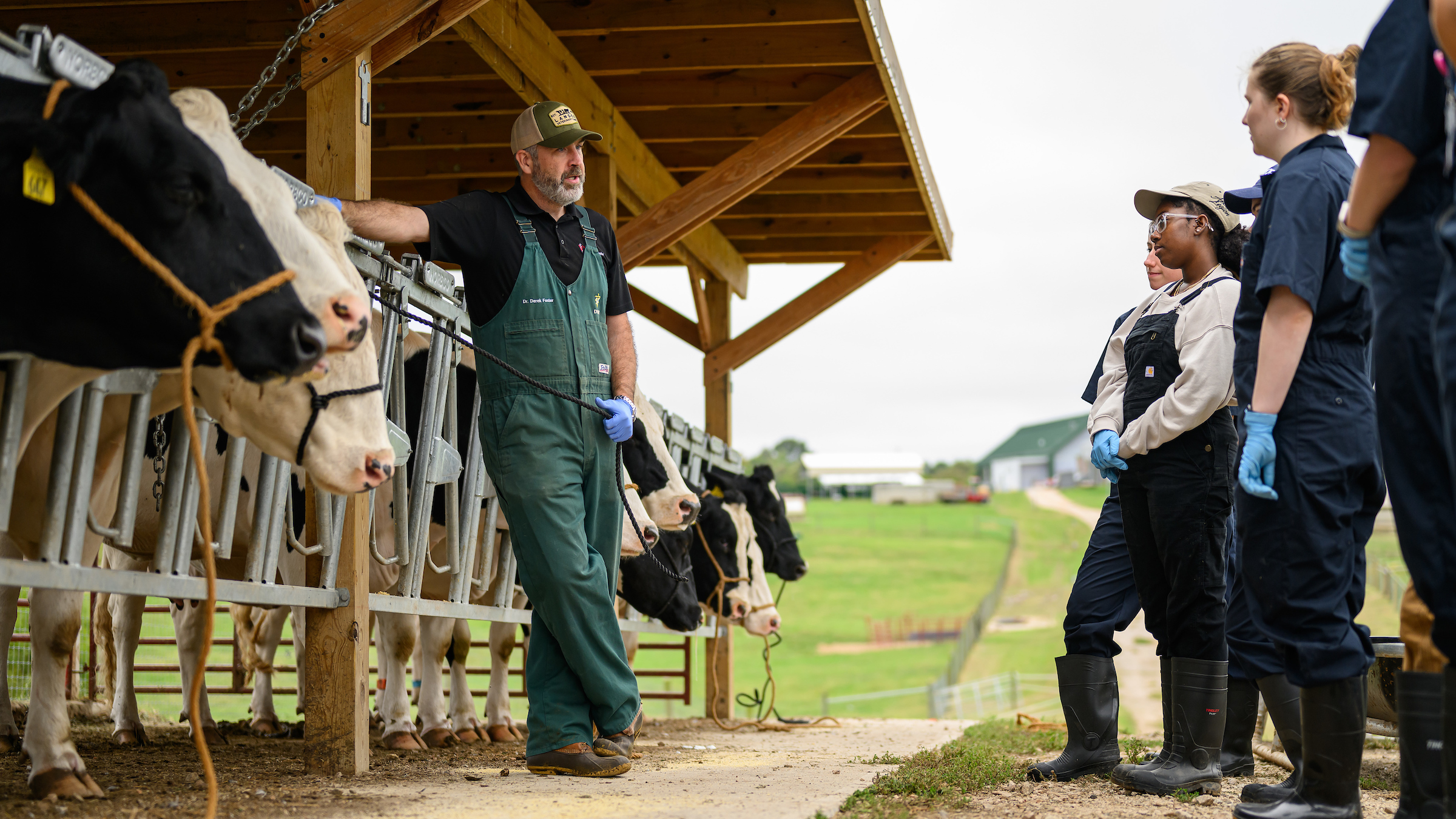 An instructor in green overalls leans against a barn containing cows while teaching a row of students in overalls during a farm lab class.