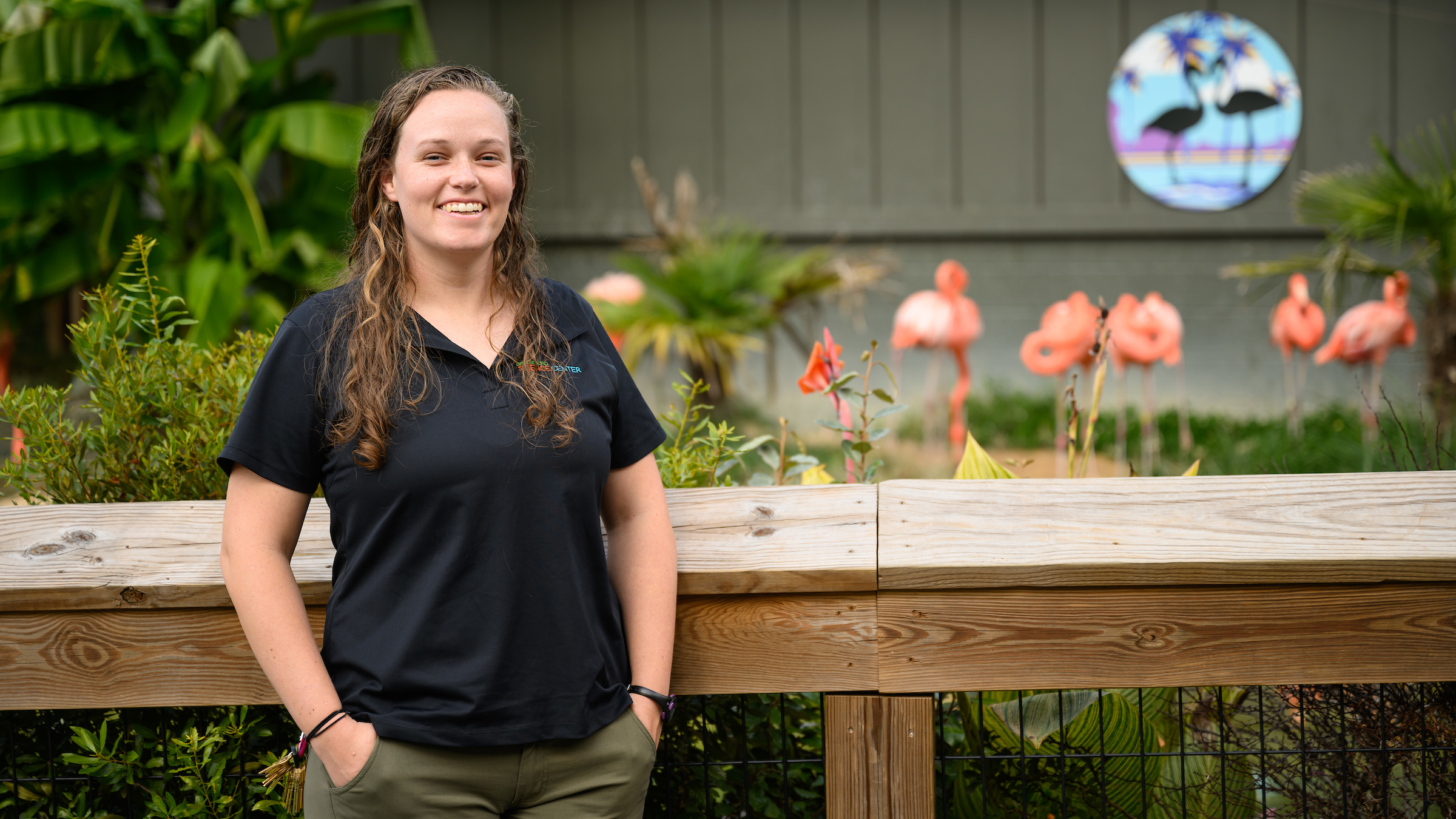 A brunette female veterinarian wearing a black shirt stands in front of a flamingo enclosure at a zoo.