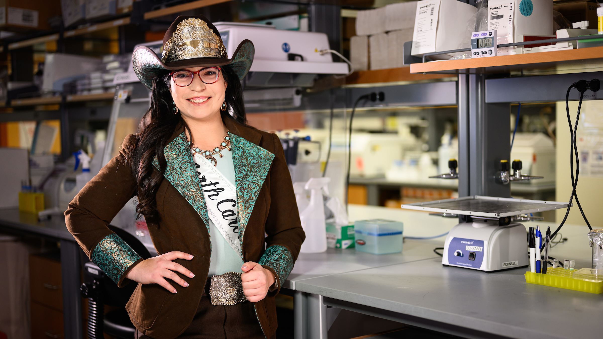 Miss Rodeo North Carolina, Rachel Gagliardi, poses in a laboratory. She has long, dark hair and glasses and is wearing a cowboy hat, a pageant sash and a brown and turquoise jacket.