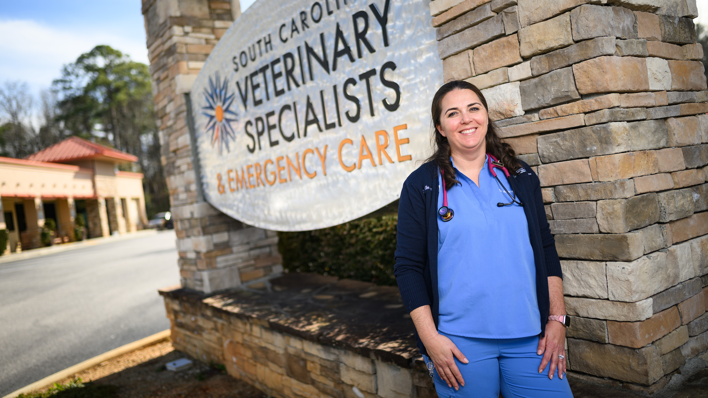 A brunette female veterinarian in light blue scrubs stands in front of the sign for her practice in South Carolina.