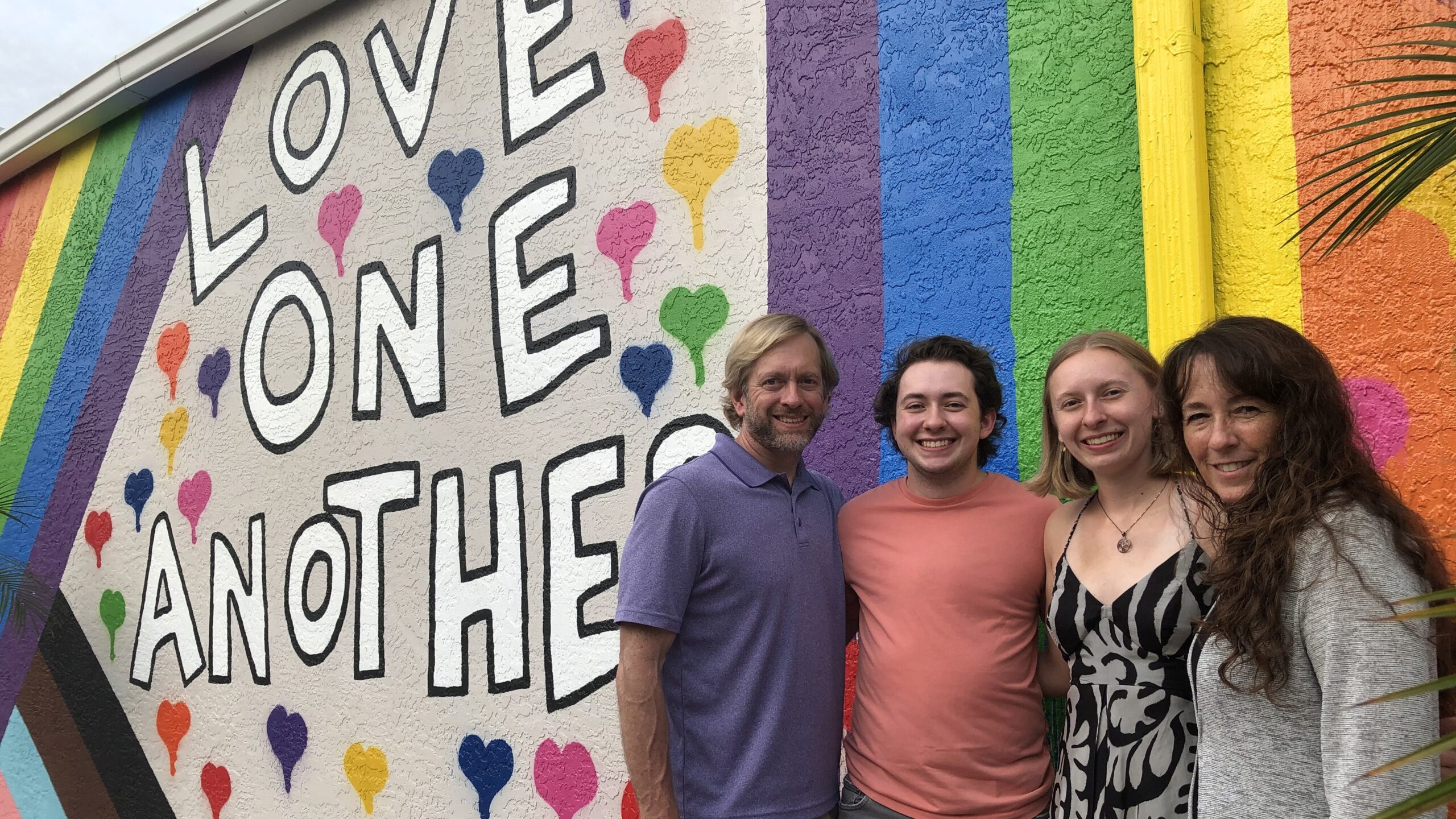 A family of four smiles in front of a rainbow mural bearing the words "Love one another." The family's father, Craig Barnett, is at the left, followed by the son, daughter and wife/mother.