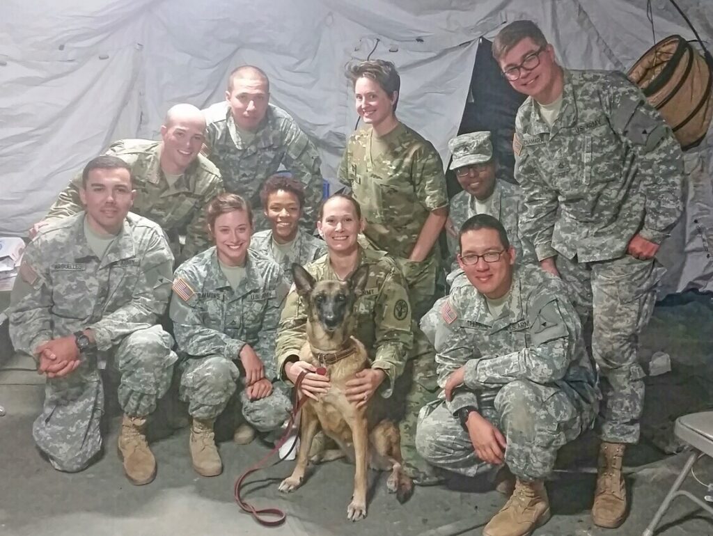 Dr. Candace Wimbish holds a German Shepherd while crouching with a team of military medics in uniform inside a white tent.