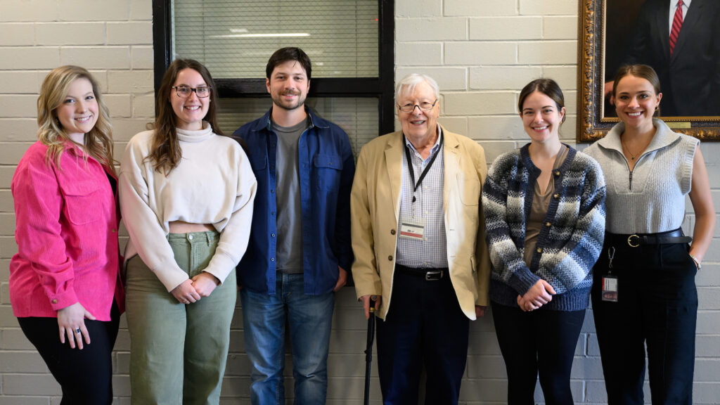 Five DVM students participating in the rural scholars program, four female and one male, stand with a former dean of the veterinary college in front of a white wall.