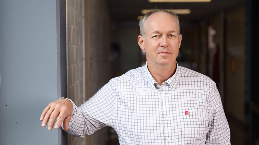 Large animal vet Dr. Jan Hawkins leans against a wall in a hospital hallway with his right hand against a windowsill. He is wearing a white shirt with a grid pattern and a small NC State logo.