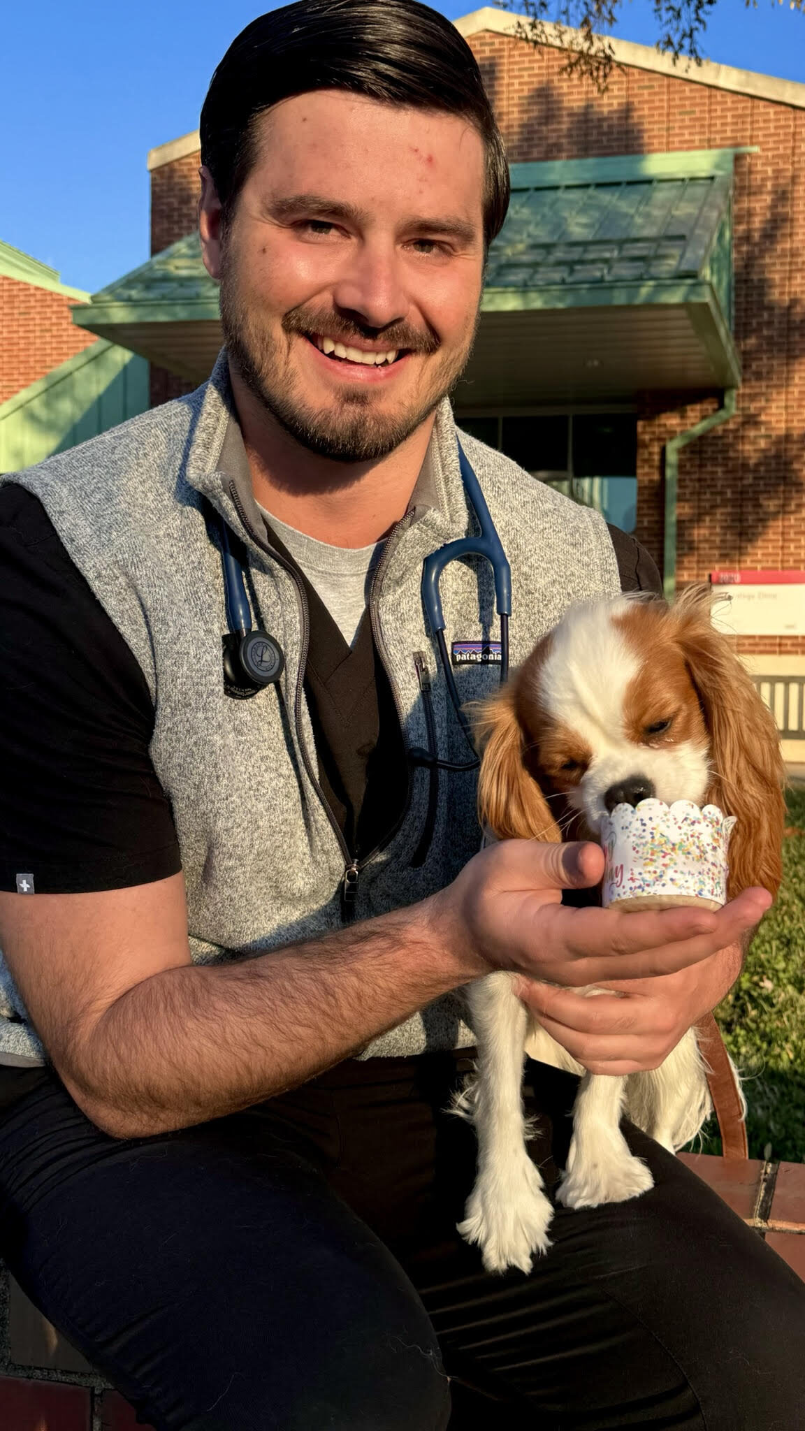 A male veterinary resident holds a Cavalier King Charles spaniel dog in front of the NC State Veterinary Hospital. The dog is licking a treat from a cup that says "Happy Birthday."