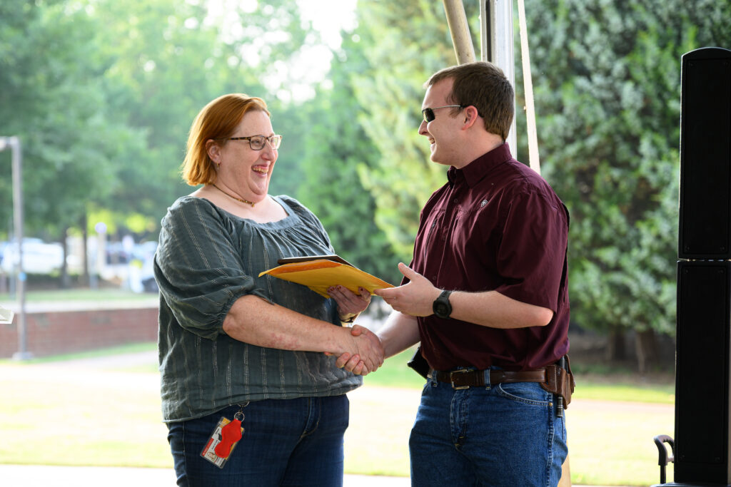 Atkinson, standing to the right, smiles as he accepts a plaque from faculty member Karen Tefft, left. Atkinson and Tefft are shaking hands as Atkinson reaches to receive the plaque from her.