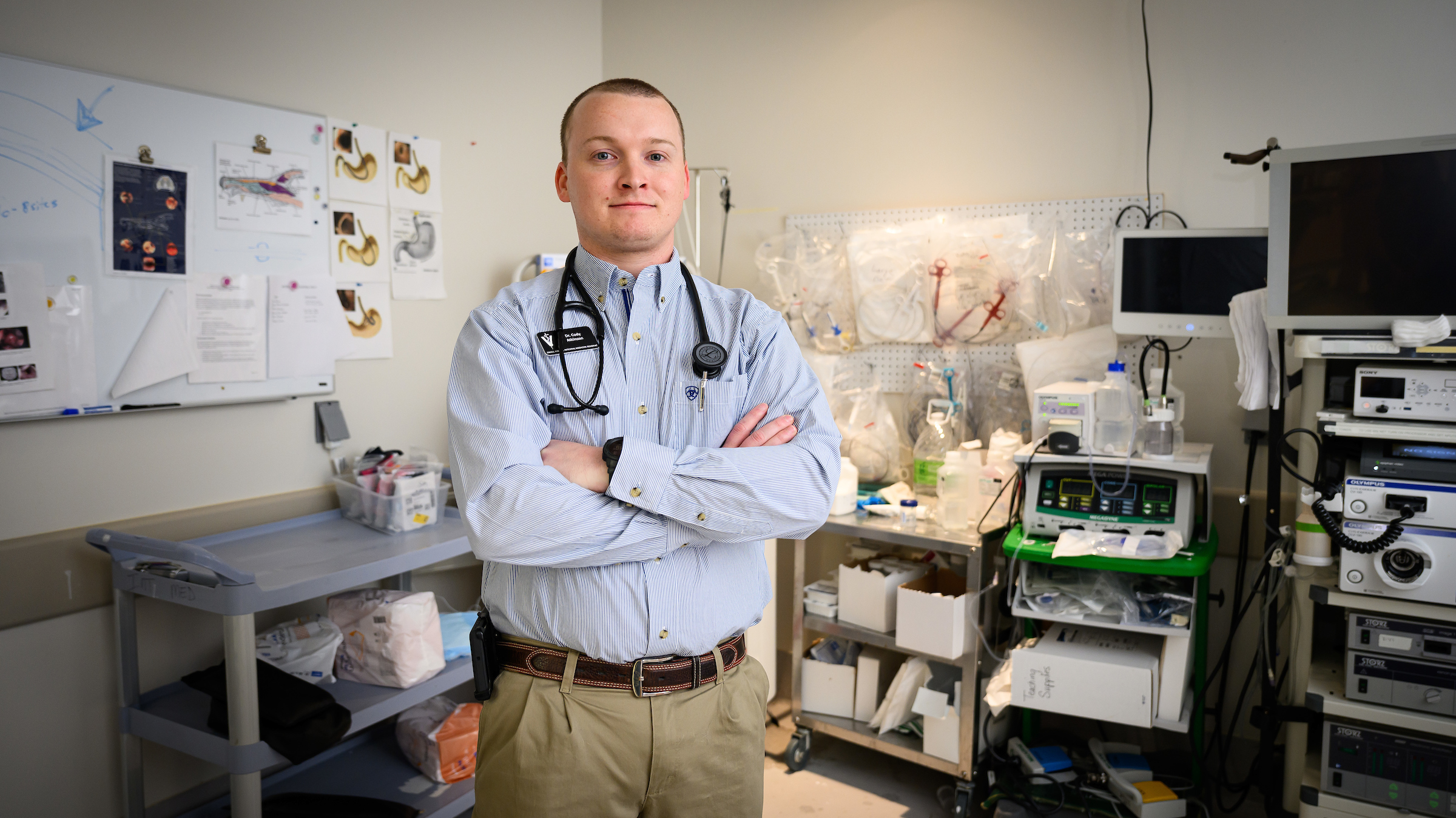 Second-year resident Dr. Cody Atkinson stands with his arms folded across his chest in front of a diagnostic room filled with medical equipment at the NC State College of Veterinary Medicine.