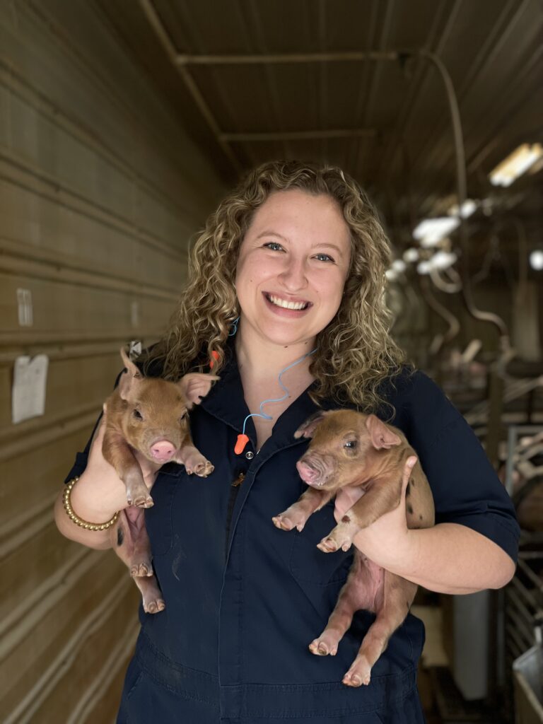 Female veterinary student Lindsey Britton smiles at the camera while holding a piglet in each hand inside a swine production facility.