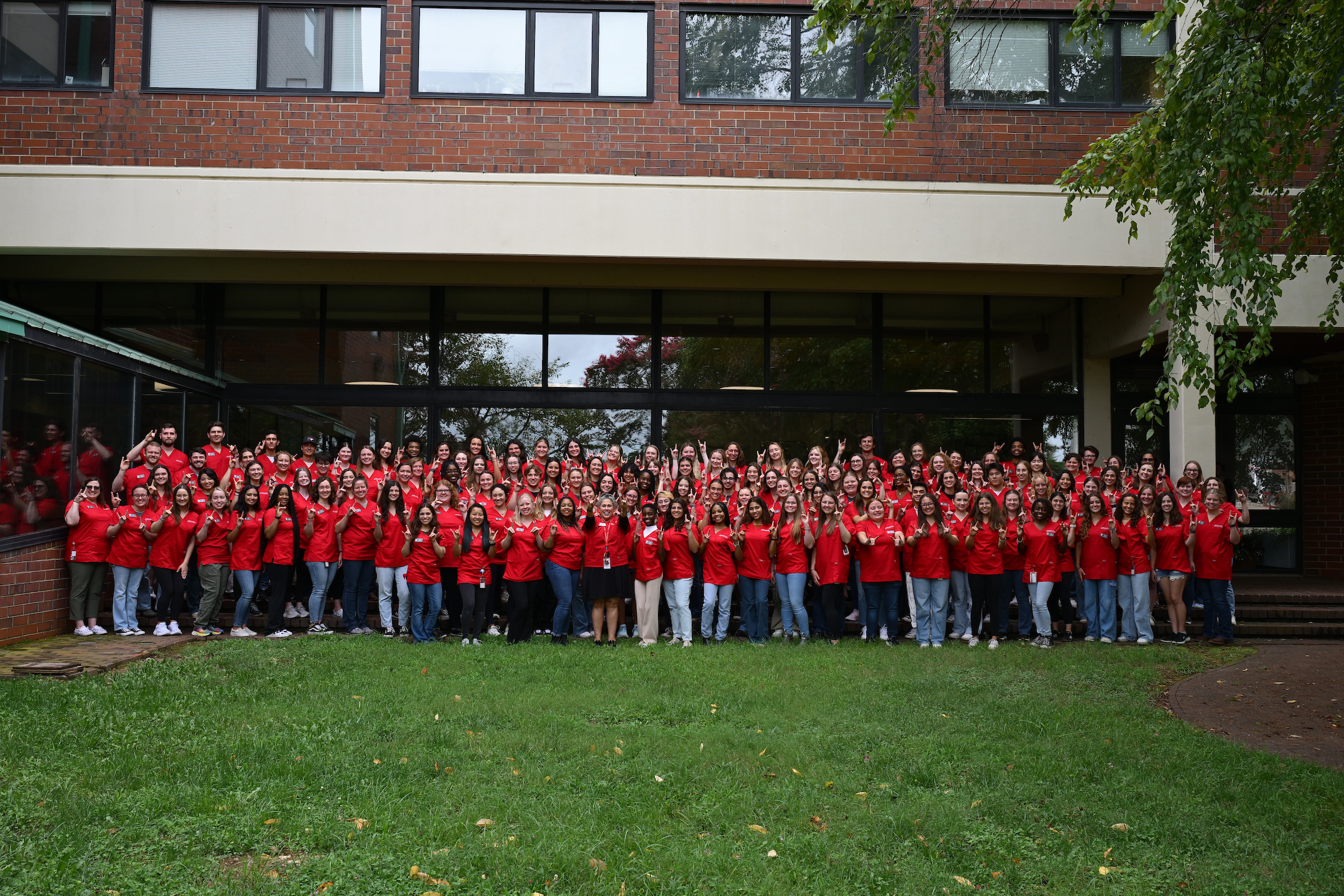 A group photo of the NC State College of Veterinary Medicine's Class of 2028 wearing red scrubs on a lawn outside the campus' main building.