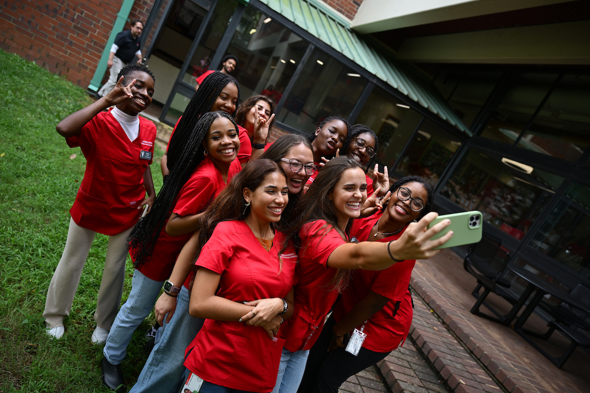 A group of female students in red scrubs smile while taking a selfie outside.