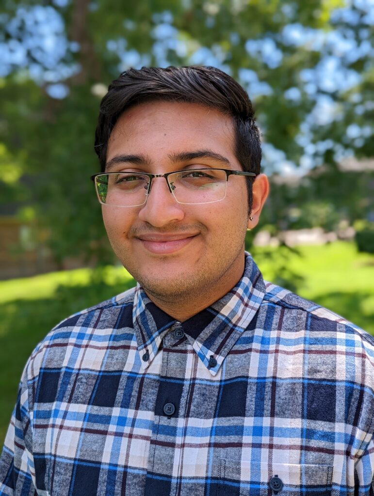 A headshot of Umang Joshi. Joshi is smiling and wearing glasses and a blue and white plaid shirt.