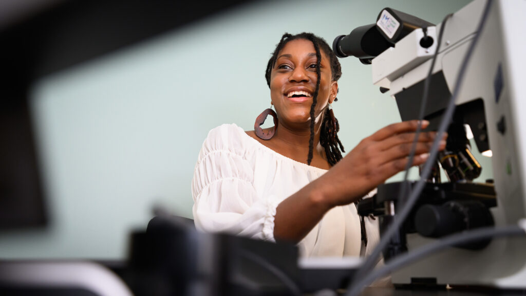 Dr. Jazz Stephens adjusts her microscope while smiling at her computer screen.