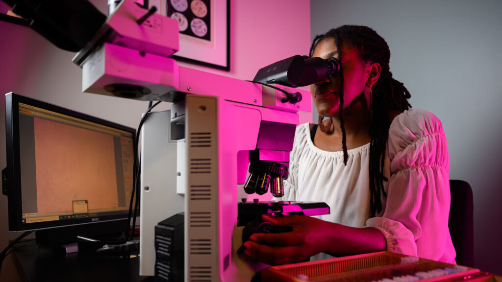 Dr. Jazz Stephens operates and peers into a microscope at a prepared slide. Her computer screen displays what she's looking at on the microscope, but all that's visible is a sea of pinkish-purple dots.