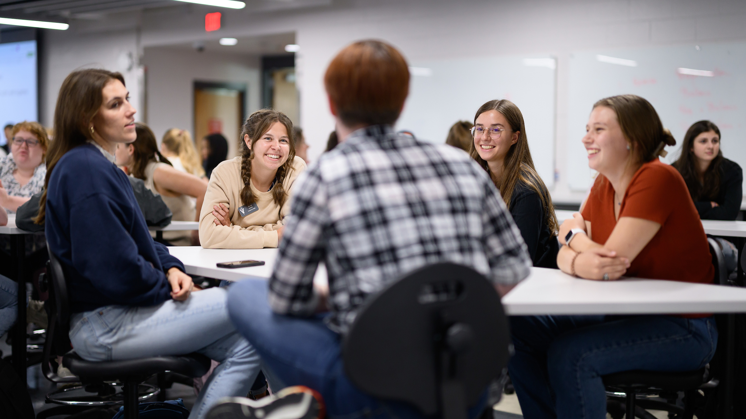 A group of five people sit around a circular table in a classroom, smiling at each other.