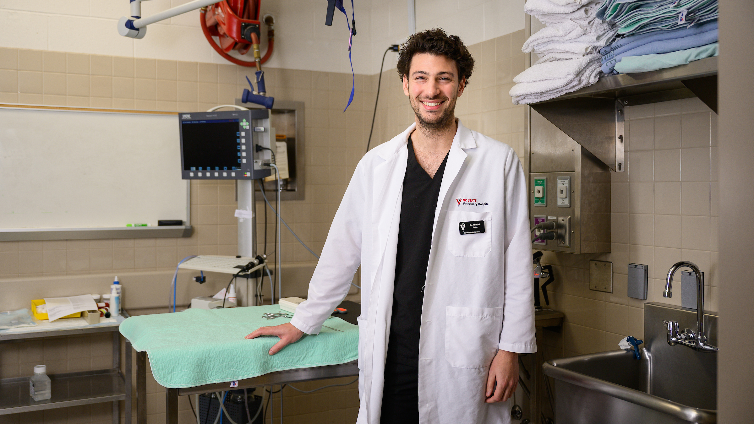 Dr. Mickael Pinto smiles at the camera while standing in a dermatology clinic at the NC State College of Veterinary Medicine. He is wearing a white veterinarian's coat and laying his hand on an examination table.