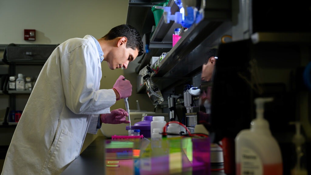 Dark-haired man in white lab coat works in a lab.