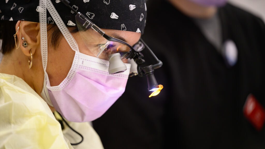 A photo zoomed in on Dr. Porcel Sánchez's masked face during an oral surgery. She is wearing a scrub cap with a molar pattern on it and wears surgical loupes.