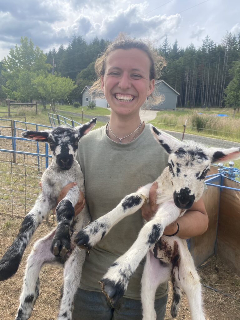 Maya Keefer stands in a livestock paddock holding a lamb under each arm.