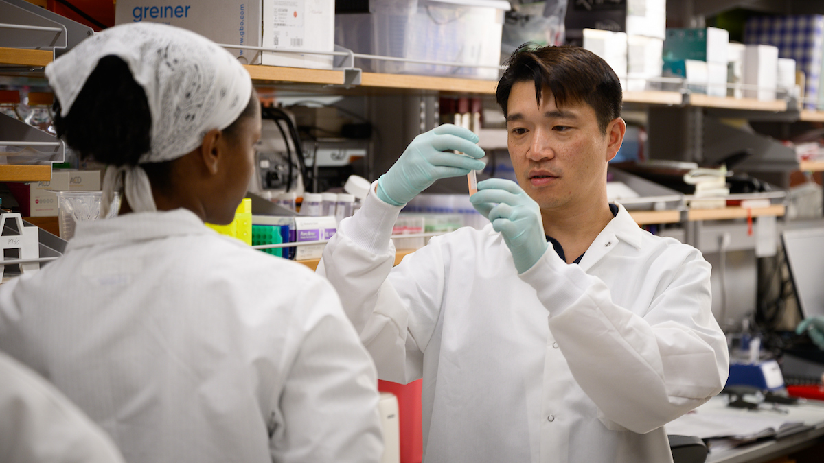 Dr. Ron Li in white coat and blue gloves looks at a vial in a lab.